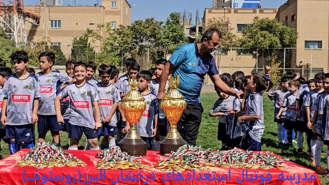 گروهی از کودکان با لباس‌های ورزشی خاکستری و آبی با آرم 'روستوف' در حال ایستادن دور یک میز قرمز که دو جام طلایی و تعداد زیادی مدال و نشان روی آن قرار دارد، همراه با یک مربی که جام‌ها را به آن‌ها اهدا می‌کند، در محیطی سبز با ساختمان‌ها در پس‌زمینه.